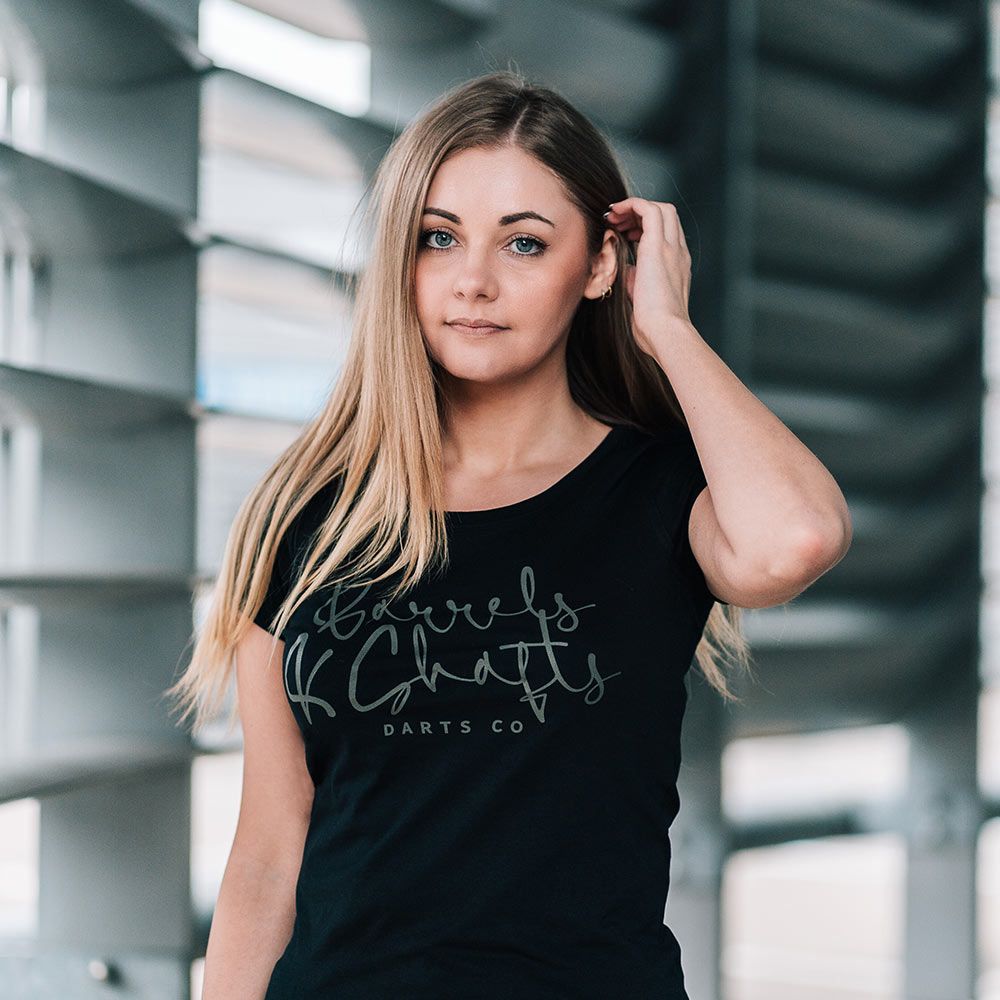 T-shirt da donna Barili e Alberi - Nera Eine Frau trägt ein schwarzes T-Shirt mit der Aufschrift „Barrels and Shafts Darts Co“. Sie steht vor einem modernen, lichtdurchfluteten Hintergrund.