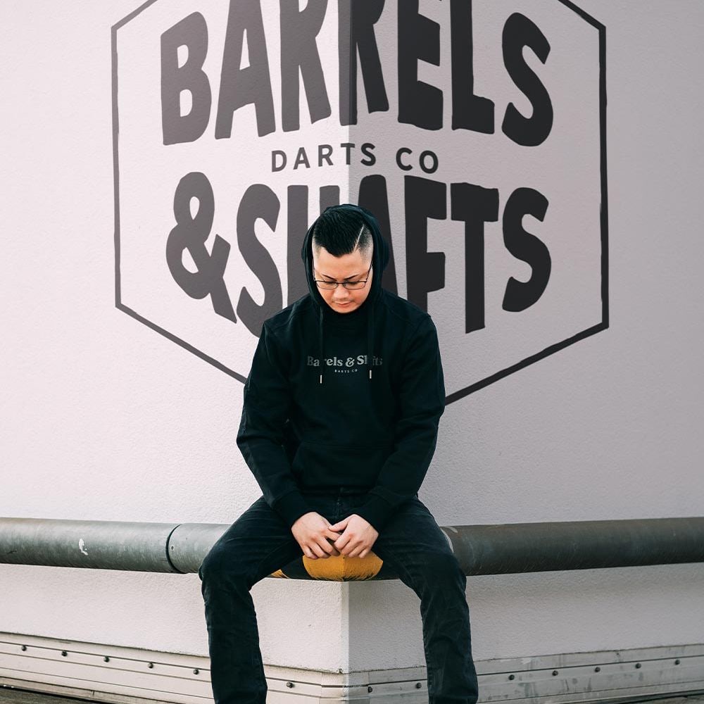 Felpa con cappuccio Barili e Alberi - Nera Eine Person trägt einen schwarzen "Barrels and Shafts" Hoodie und sitzt auf einer Bank. Im Hintergrund ist ein großes Wandlogo mit der Aufschrift "Barrels & Shafts Darts Co" zu sehen.
