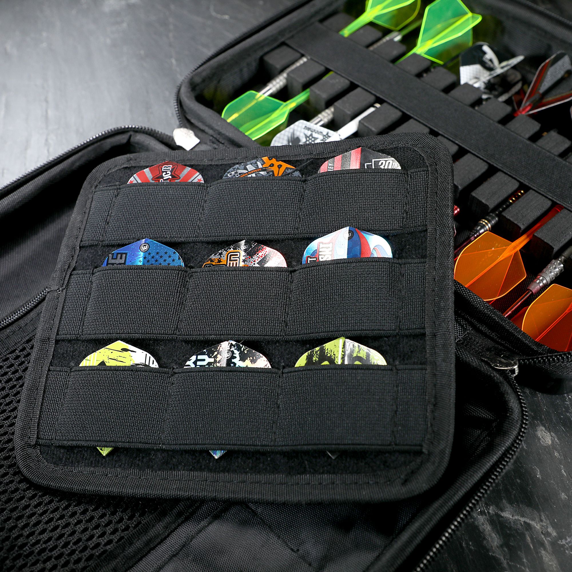 Winmau Armor G4 Dartwallet In dem Bild sieht man ein schwarzes Dartetui, das verschiedene Darts und Dart-Flights ordentlich aufbewahrt. Die Darts sind auf der rechten Seite einsortiert, während links viele bunte Flights in Fächern stecken.