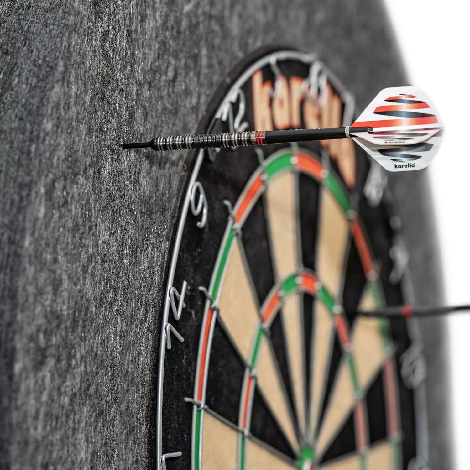 Cornice per bersaglio in acciaio Karella Ultimo Das Bild zeigt eine Karella Ultimo Steeldartboard Surround, die einen Dartpfeil auffängt. Die graue Umrandung schützt die Wand vor Fehltreffern neben der Dartscheibe.