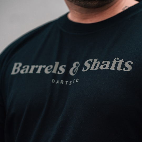 T-shirt barili e alberi - nera Das Bild zeigt ein schwarzes T-Shirt mit der Aufschrift "Barrels & Shafts". Darunter steht in kleineren Buchstaben "DARTSCO".