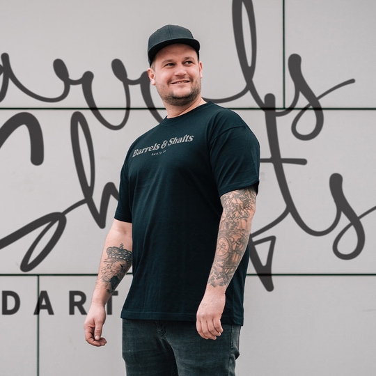T-shirt barili e alberi - nera Ein Mann trägt ein schwarzes T-Shirt mit der Aufschrift „Barrels & Shafts“. Er steht vor einer Wand mit passender Schrift im Hintergrund.