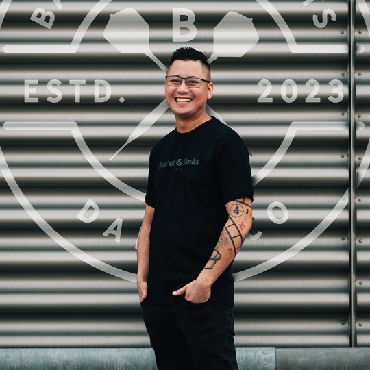 T-shirt barili e alberi - nera Ein Mann trägt ein schwarzes "Barrels and Shafts"-T-Shirt und steht vor einer metallenen Wand mit großem Logo. Er lächelt und hat markante Tattoos am rechten Arm.