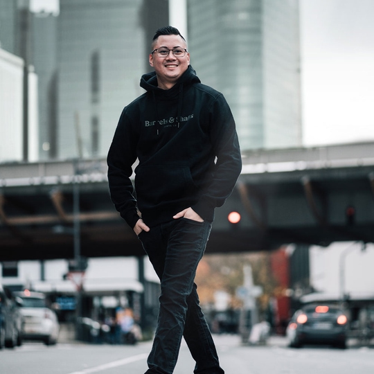 Felpa con cappuccio Barili e Alberi - Nera Eine Person trägt einen schwarzen "Barrels and Shafts" Hoodie und steht entspannt auf einer Straße. Im Hintergrund sind unscharfe Gebäude und Autos zu sehen.