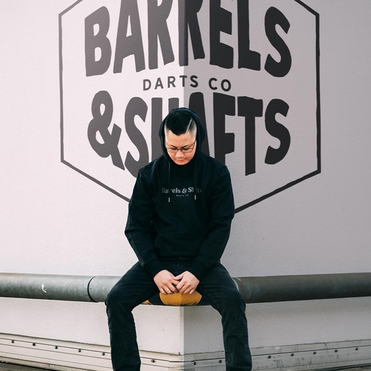 Felpa con cappuccio Barili e Alberi - Nera Eine Person trägt einen schwarzen "Barrels and Shafts" Hoodie und sitzt auf einer Bank. Im Hintergrund ist ein großes Wandlogo mit der Aufschrift "Barrels & Shafts Darts Co" zu sehen.