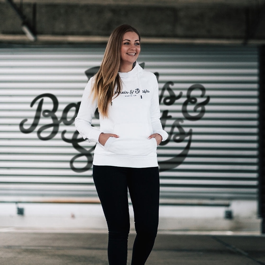 Felpa con cappuccio da donna Barrels and Shafts - bianca Eine Frau trägt einen weißen "Barrels and Shafts" Damen Hoodie. Sie steht vor einem Hintergrund mit großem Schriftzug und lächelt.